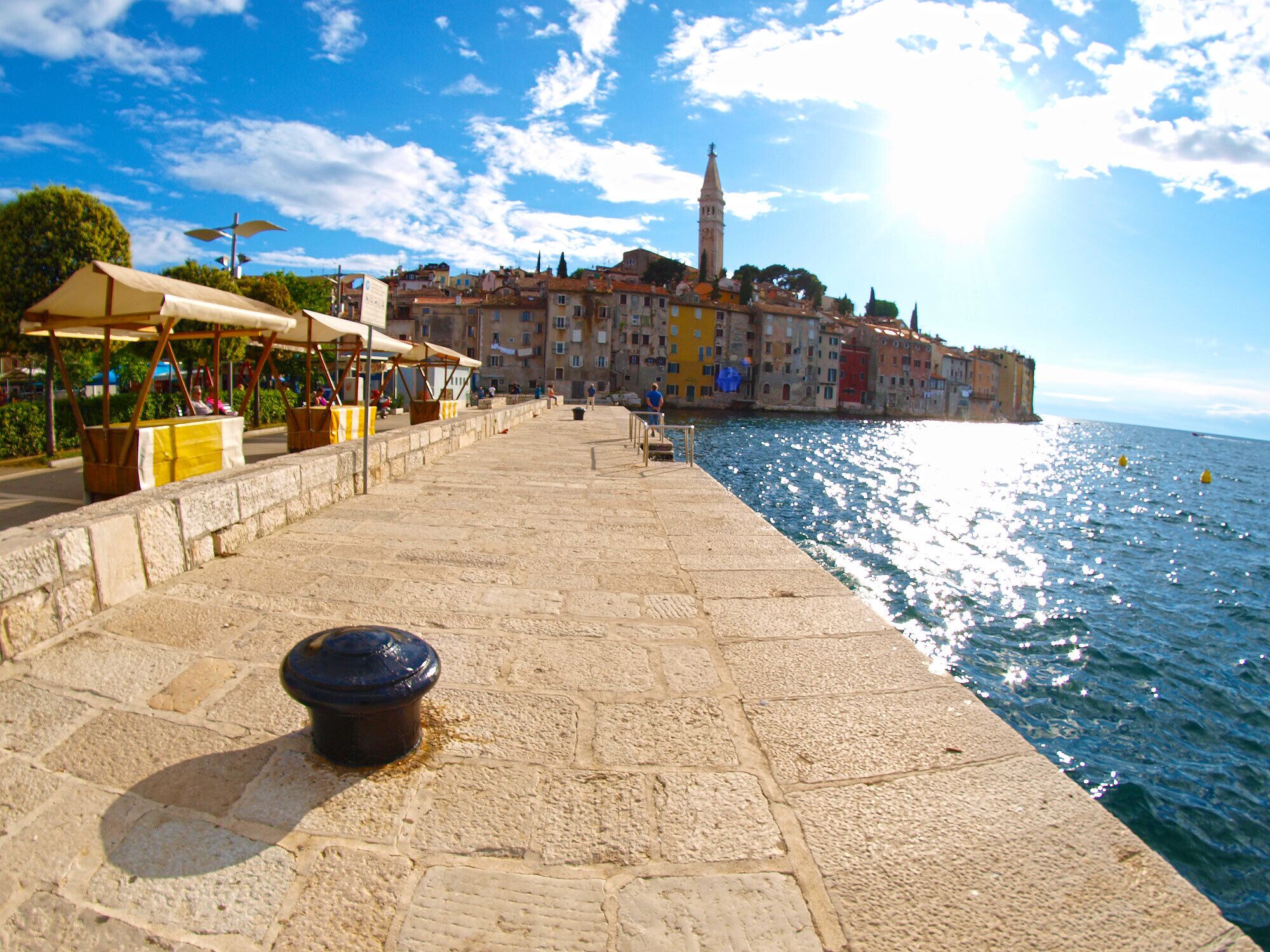 A stone-paved waterfront promenade with market stalls and a historic building in the background.
