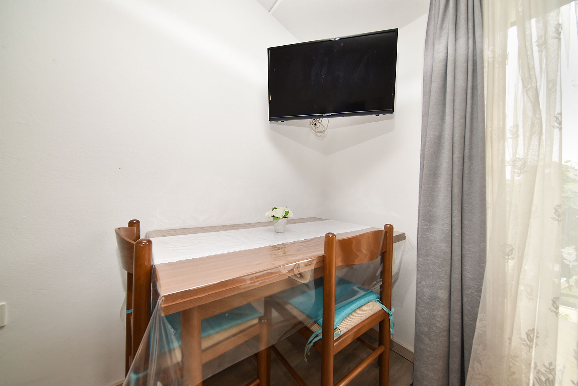 A small room with a wooden table, two chairs, a flat-screen TV mounted on the wall, and a window with curtains.