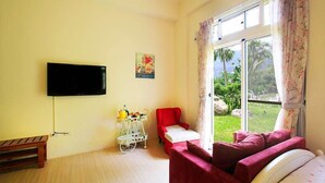 A room with a TV mounted on the wall, a red chair, a small table with a white chair, and a window with curtains. - Provence Villa (Nanzhuang)