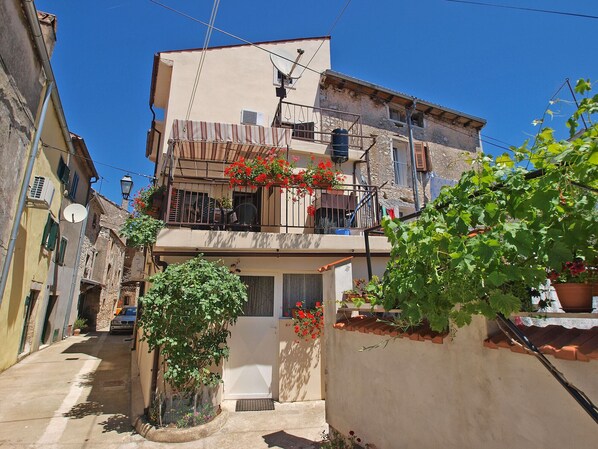A narrow street with buildings on either side, a small tree in the foreground, and a balcony with red flowers. - Apartment 1123/9662 (Istria - Vodnjan), 5000m from the beach (Vodnjan)