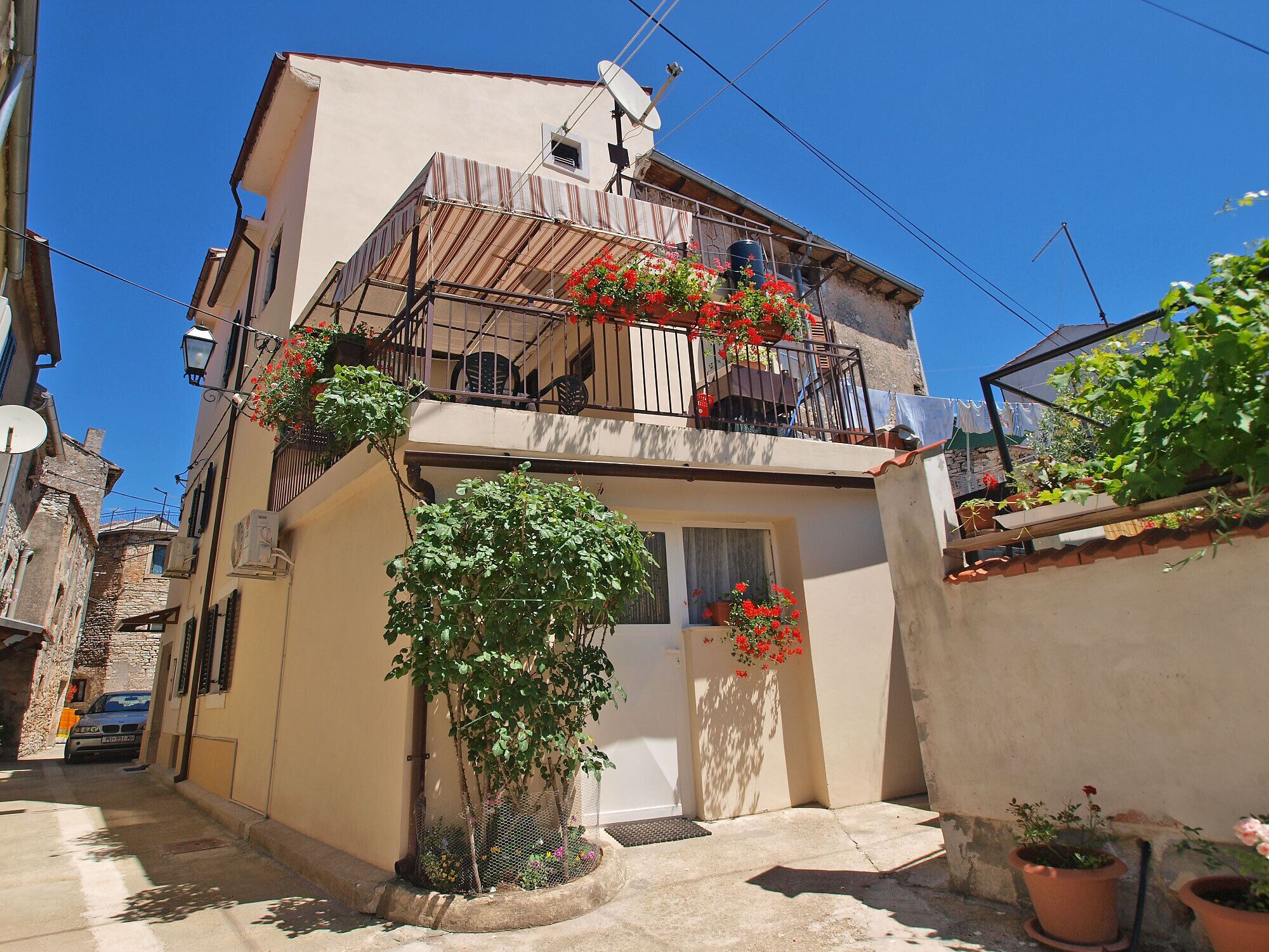 A narrow street with a building featuring a balcony with red flowers, a small garden, and a satellite dish.