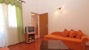 Ein Wohnzimmer mit Holzboden, einem Glastisch, einem orangefarbenen Sofa mit Kissen, einem Fernseher auf einem Holzschrank und einem grünen Vorhang.