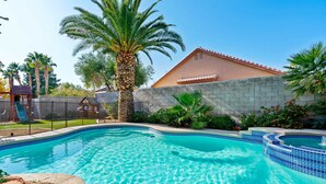A swimming pool with a stone wall, palm trees, and a house in the background. - LAS VEGAS LUXURY HOME, 5 BDRP, POOL W/HEATER, BBQ, GAMES (Las Vegas)