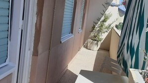 A narrow outdoor corridor with a white table and chairs, a small potted plant, and a window with closed shutters.