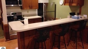 A kitchen with wooden cabinets, a stainless steel oven, a refrigerator, and a bar area with stools. - Crystal Mtn Condo, walk to lift,  soaking tub, short drive to Iron Fish & Lk MI (Thompsonville)