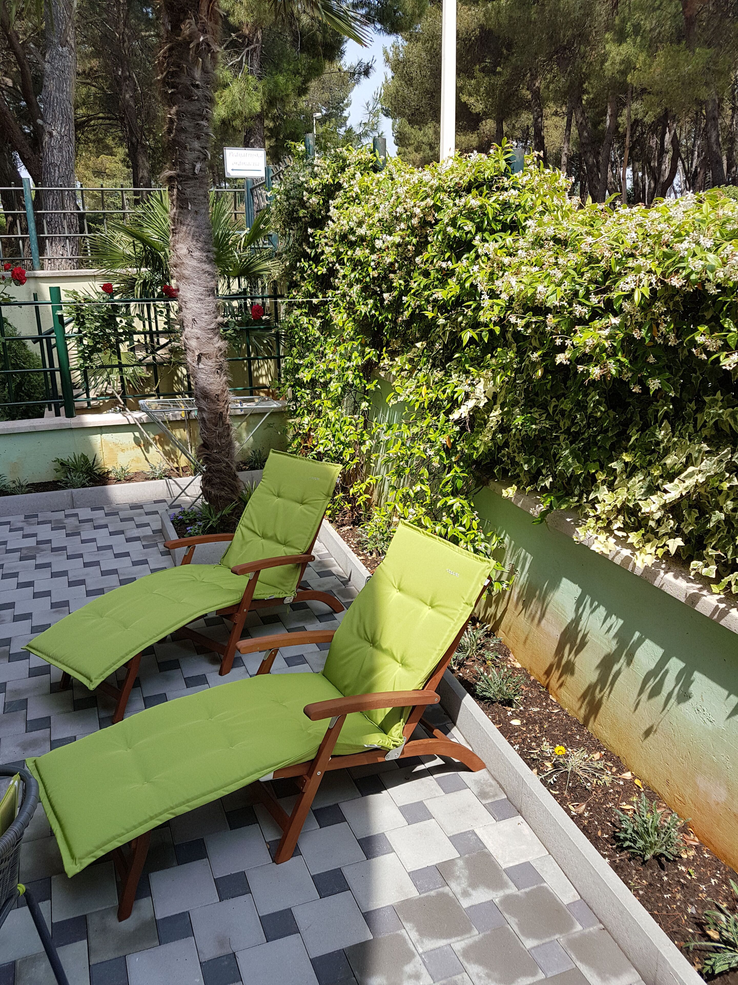 Two green lounge chairs on a tiled patio with a hedge and trees in the background.
