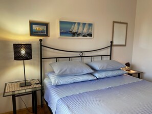 A bedroom with a bed, two pillows, a nightstand with a lamp, a framed painting, and a mirror.