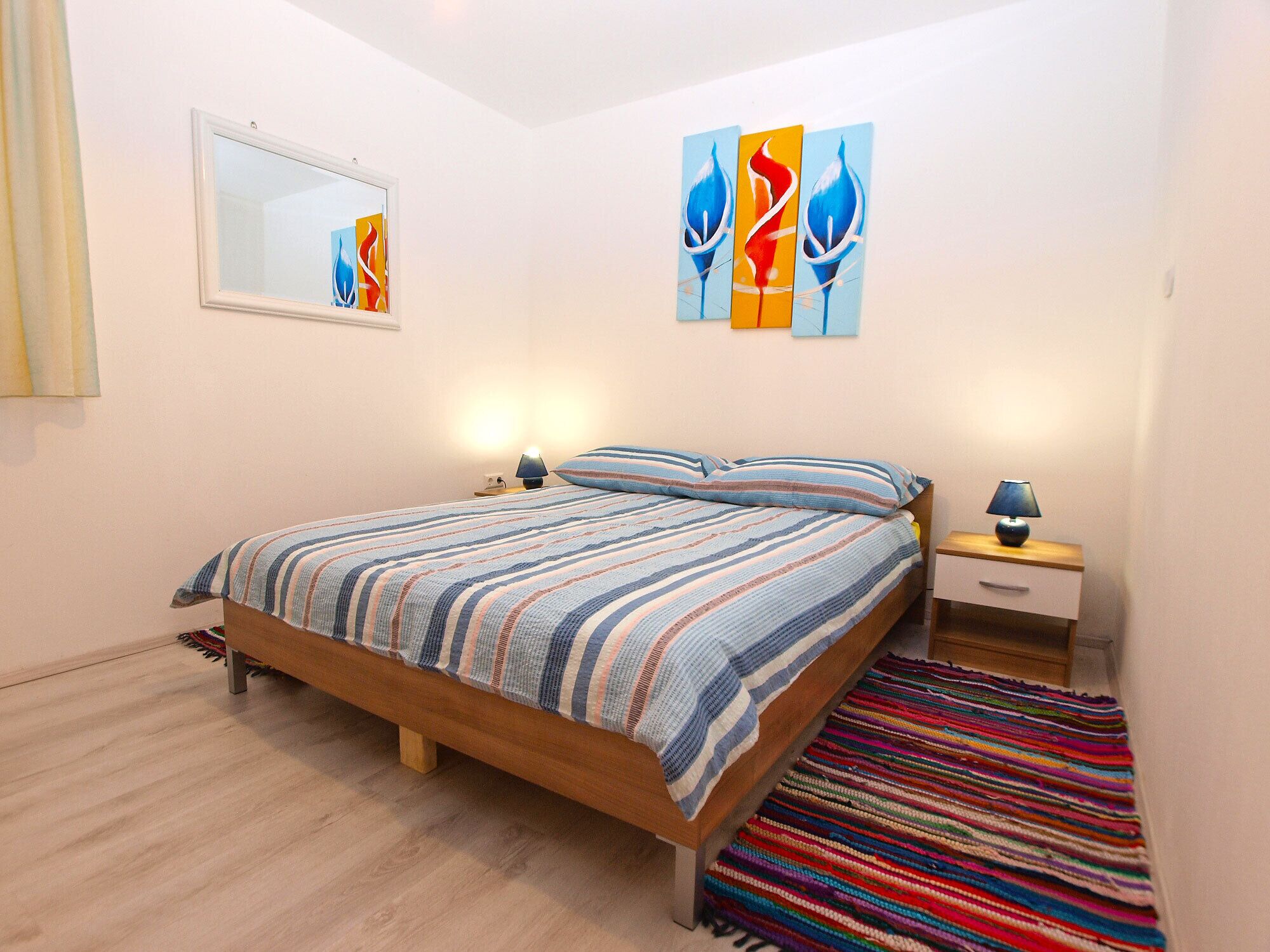 A bedroom with a wooden bed, two bedside tables with lamps, a colorful striped rug, and abstract wall art.