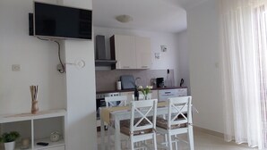 Une cuisine compacte avec des meubles blancs, un coin repas avec des chaises blanches, un téléviseur fixé au mur et une petite étagère ornée d’une plante et d’une horloge.