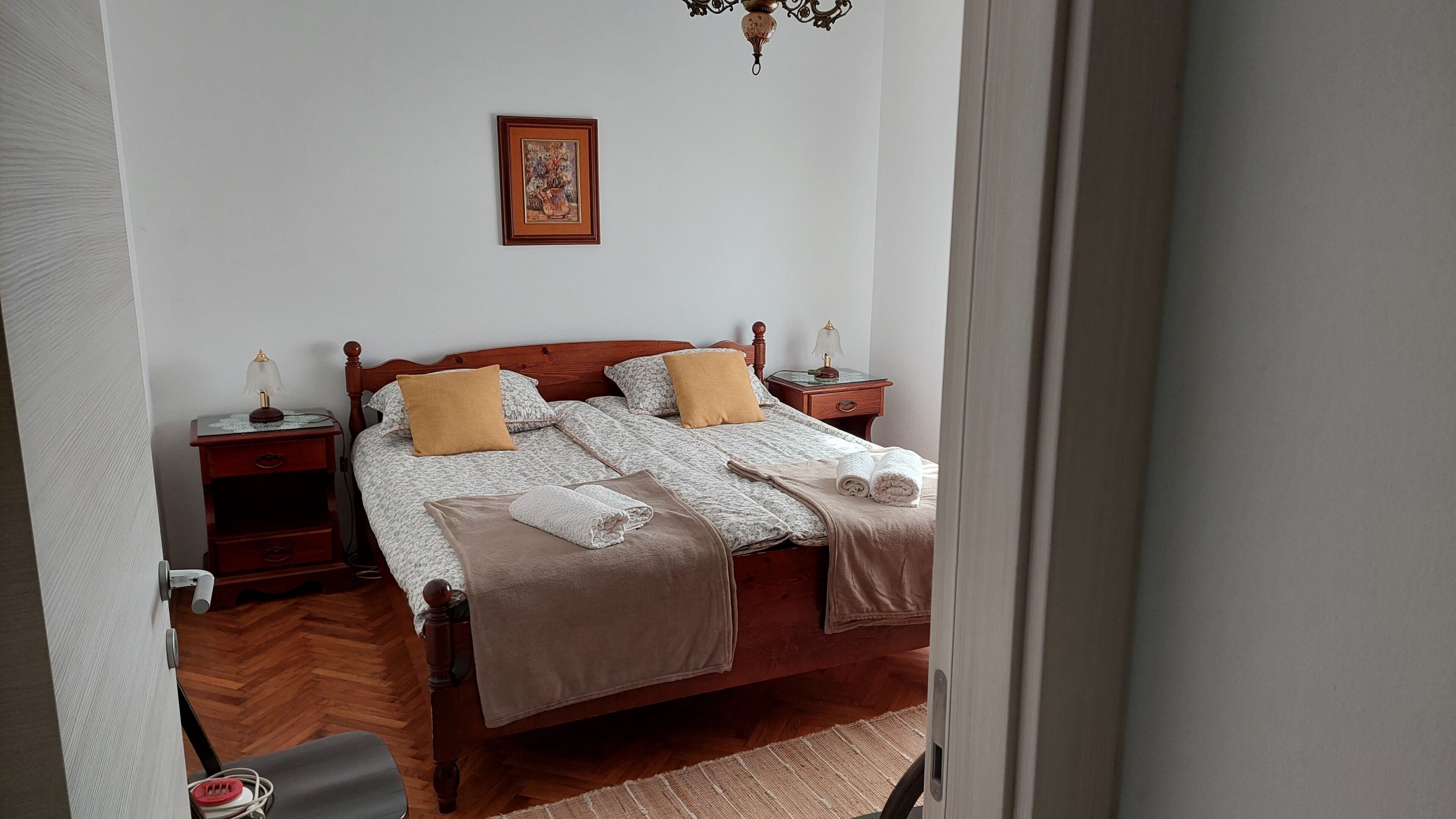A neatly made bed with a wooden headboard, two bedside tables with lamps, a framed picture on the wall, and a wooden floor.