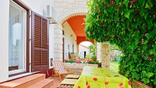 A patio area with a table covered in a yellow tablecloth, stone steps leading to a door, and a view of a garden through an archway. - Apartment 1189/10858 (Istria - Premantura), 500m from the beach (Premantura)