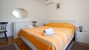 A bedroom with a bed, a chair, a bedside table, a lamp, a mirror, and an air conditioning unit.