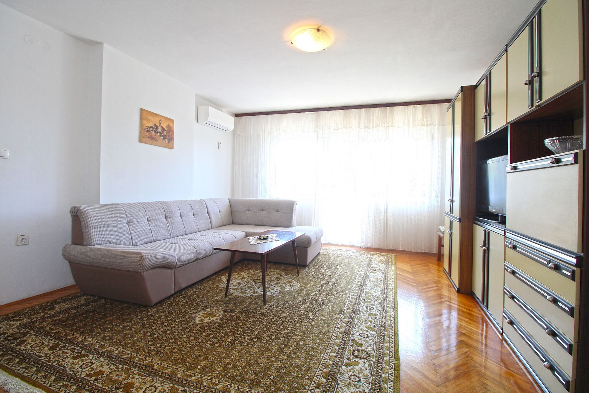 A living room with a sofa, a coffee table, a patterned rug, and a kitchen area with cabinets and a refrigerator.