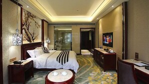 A hotel room with a large bed, bedside tables, a TV, a desk, and a bathroom visible through a glass door. - Jin Long Wan Hao Hotel (Wuzhou)