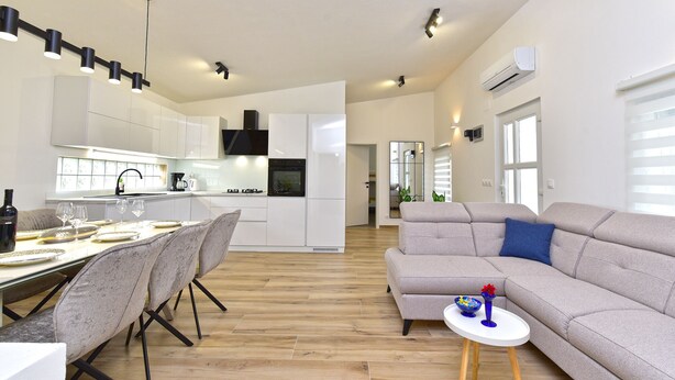 A modern kitchen with white cabinets, a dining area with a table and chairs, a grey sofa, and a small side table with decorative items.
