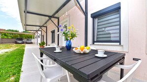 An outdoor patio with a table set for two, white chairs, a vase with flowers, and a bowl of fruit.