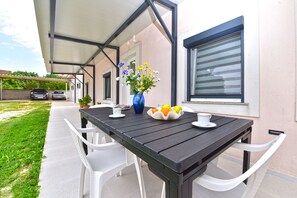 An outdoor patio with a wooden table, white chairs, a vase with flowers, and a bowl of fruit.