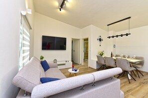 A modern living room with a sofa, a dining area with a table and chairs, a TV mounted on the wall, and a ceiling light fixture.
