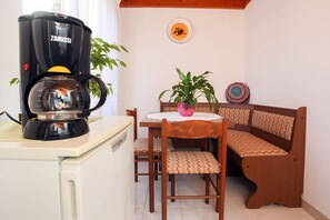 Uma cozinha compacta com cafeteira Zanussi, geladeira branca, mesa de jantar de madeira com uma planta rosa e um banco de madeira com almofadas estampadas.