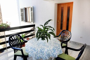 Uma pequena área de estar ao ar livre, com uma mesa coberta por toalha branca estampada em azul, duas cadeiras pretas com almofadas verdes e um vaso de planta.
