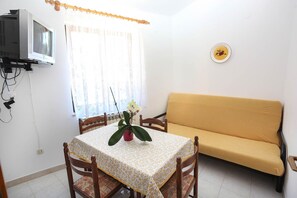 Uma pequena área de refeições com uma mesa coberta por um guardanapo estampado, duas cadeiras de madeira, um sofá amarelo e uma TV fixada na parede.