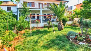 A two-story house with a balcony, surrounded by a garden with various plants and a palm tree.