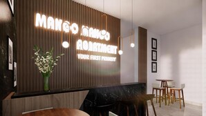 Un moderno mostrador de recepción con un panel de madera y un letrero de neón que dice “Mango Mango Apartment. Your First Priority.” Hay cuadros enmarcados en la pared, un florero con flores y un área de descanso con sillas y una mesa.