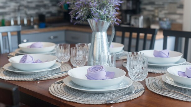 Una mesa de comedor dispuesta con platos, tazones y cubiertos blancos, acompañada de copas transparentes y un florero con flores moradas.