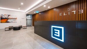 Un espace d’accueil au design moderne, avec un mur revêtu de panneaux en bois sur lequel figure le mot « ELITE » et un logo sur la droite. On y trouve également des coins salon avec des poufs ronds et un panneau de sécurité incendie sur la gauche.