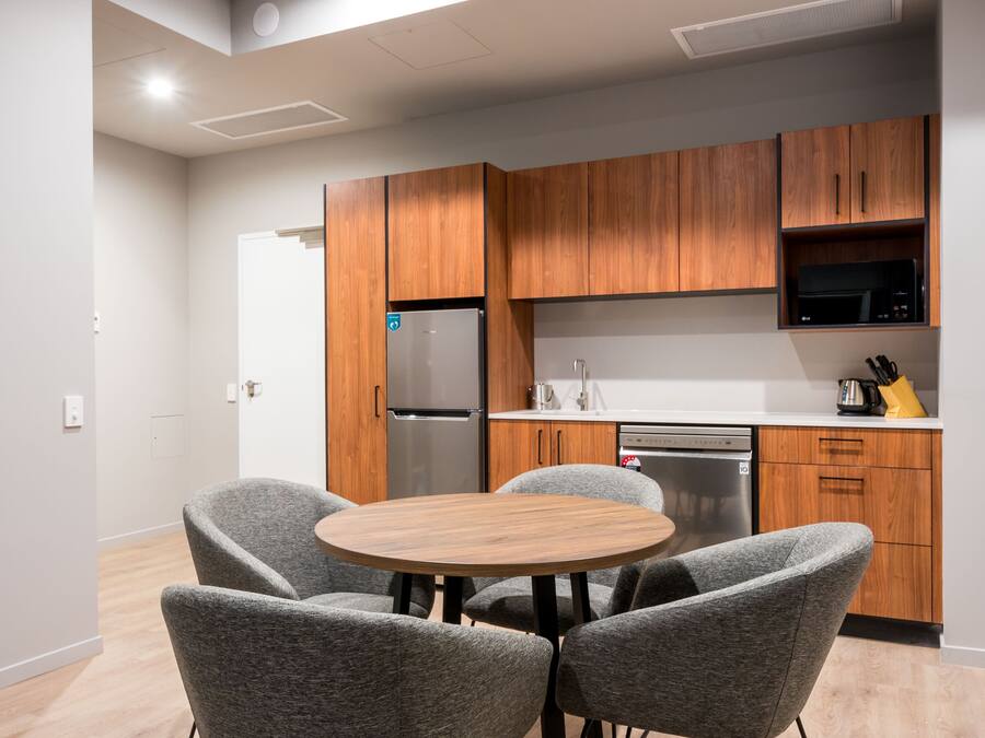A modern kitchen with wooden cabinets, a stainless steel refrigerator, and a built-in microwave. There is a round wooden table with four grey chairs.