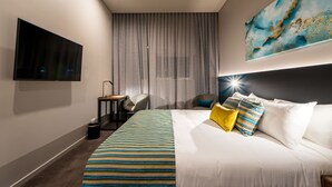 Une chambre d’hôtel moderne dotée d’un grand lit, d’un téléviseur à écran plat fixé au mur, d’un bureau avec une chaise et d’une grande fenêtre avec des rideaux.