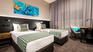 A hotel room with two beds, a desk, a chair, a large abstract painting on the wall, and a window with curtains.