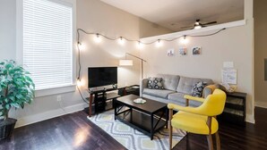 A living room with a sofa, a yellow armchair, a coffee table, a TV, and string lights. - Modern & Stylish 1BR ◈ Great Location ◈ Walkable (Charlotte)