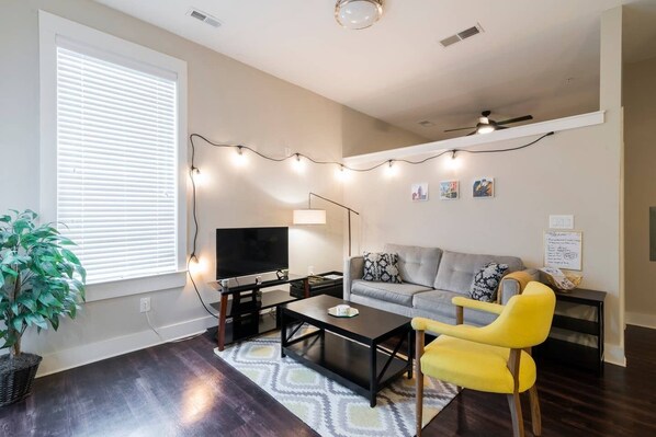 A living room with a sofa, a yellow armchair, a coffee table, a TV, and string lights.