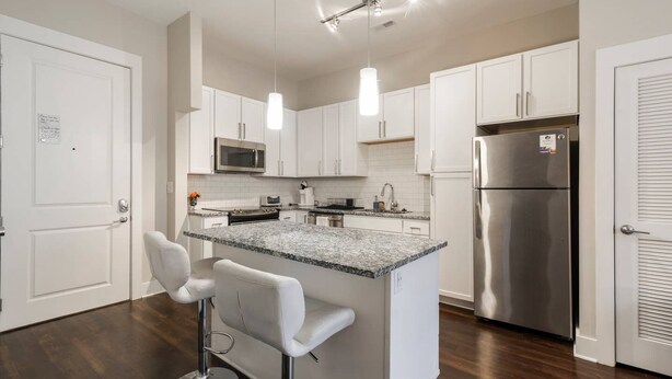 A modern kitchen with white cabinets, a stainless steel refrigerator, a microwave, and a granite countertop island with bar stools. - Modern & Stylish 1BR ◈ Great Location ◈ Walkable (Charlotte)