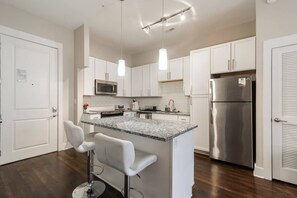 A modern kitchen with white cabinets, a stainless steel refrigerator, a microwave, and a granite countertop island with bar stools.