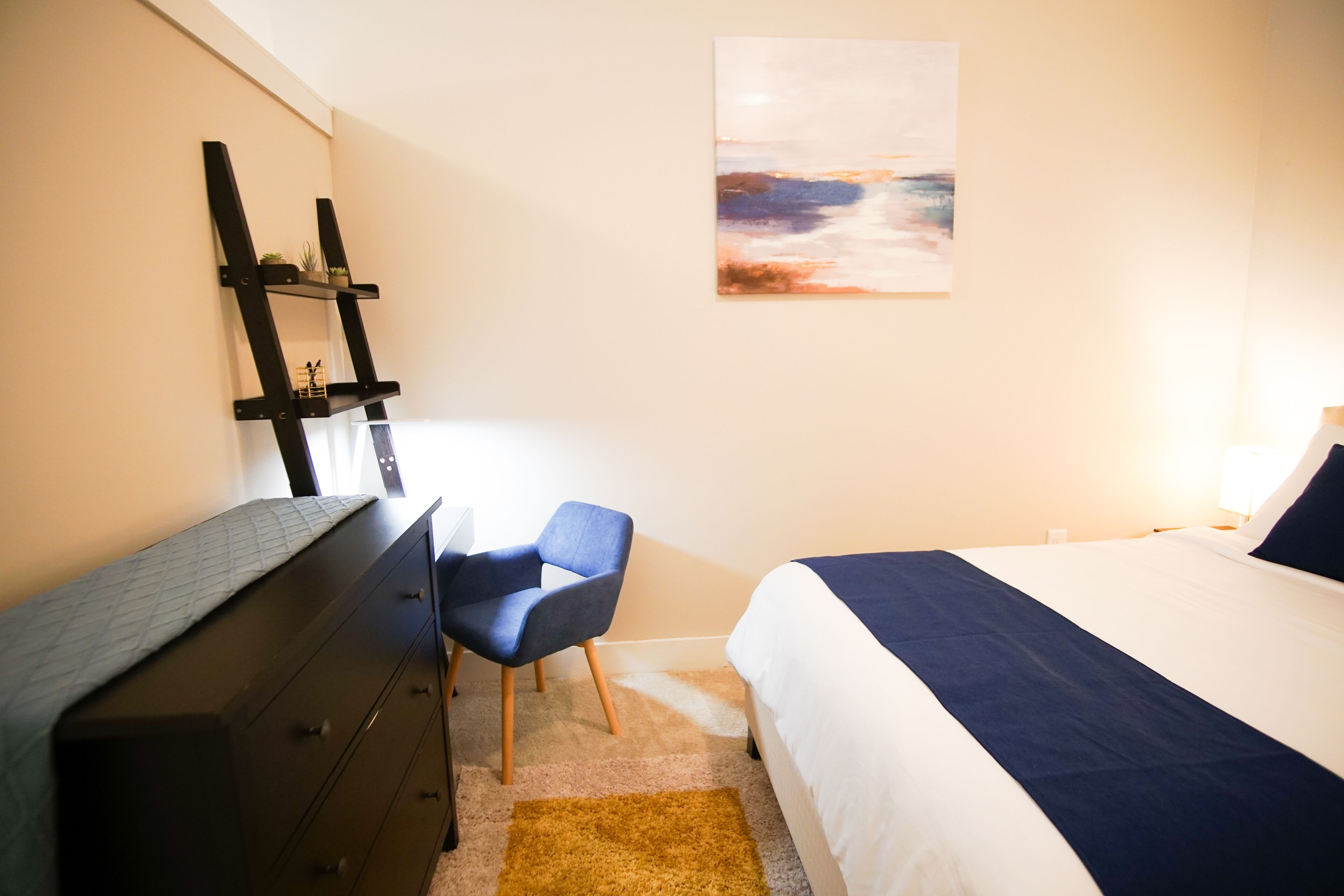 Un dormitorio con cama, escritorio, silla, cómoda y un cuadro en la pared.