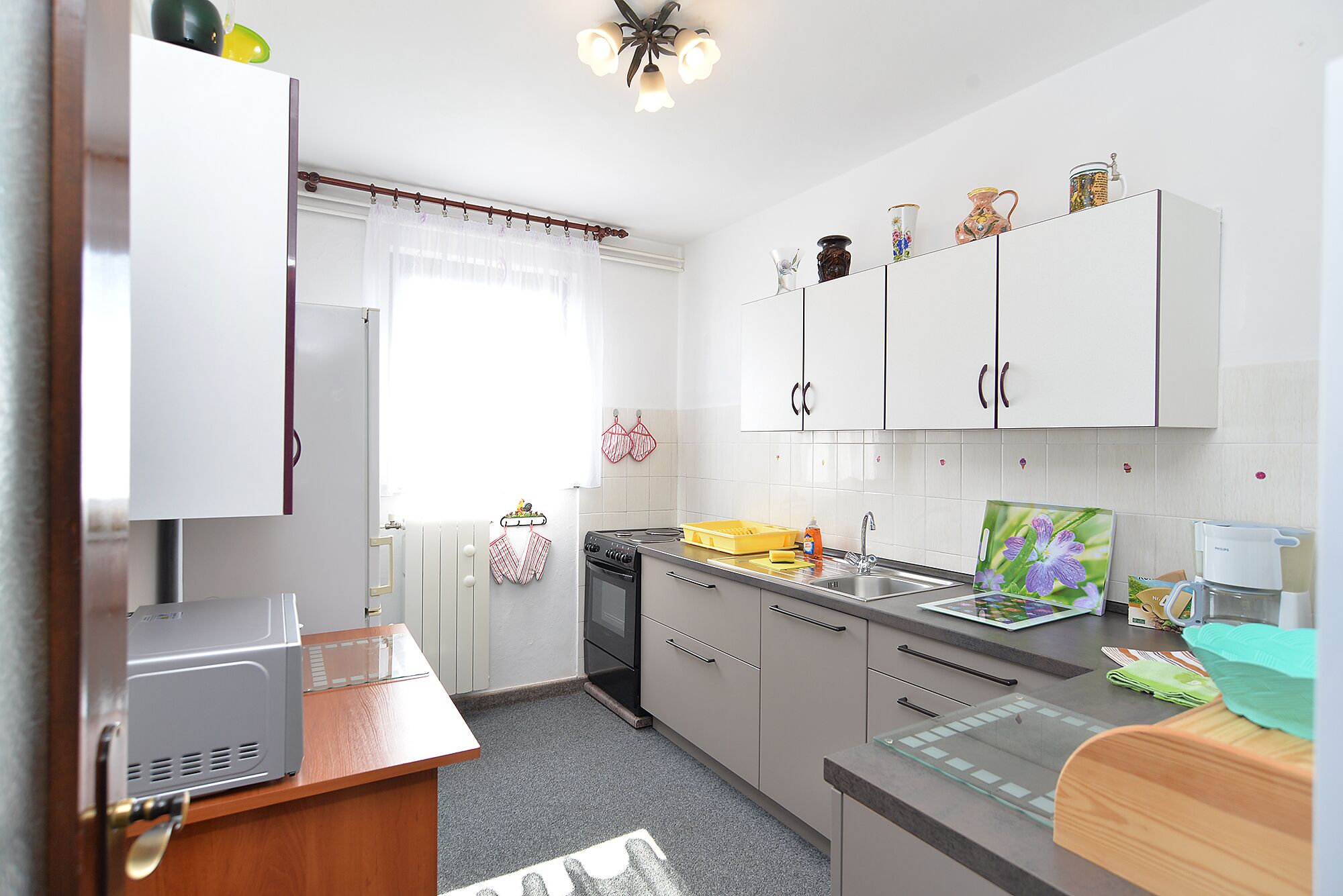 A modern kitchen with white cabinets, a stove, and a sink. There is a window with curtains, a radiator, and a small table with a toaster.