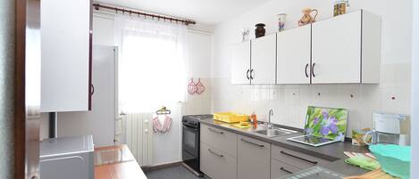 A modern kitchen with white cabinets, a stove, and a sink. There is a window with curtains, a radiator, and a small table with a toaster.