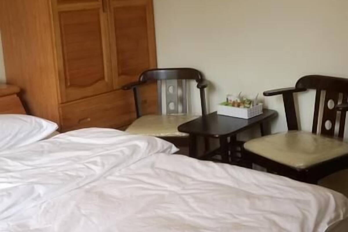 A room with a large wooden wardrobe, two wooden chairs, a small table with a basket, and a bed with white bedding.
