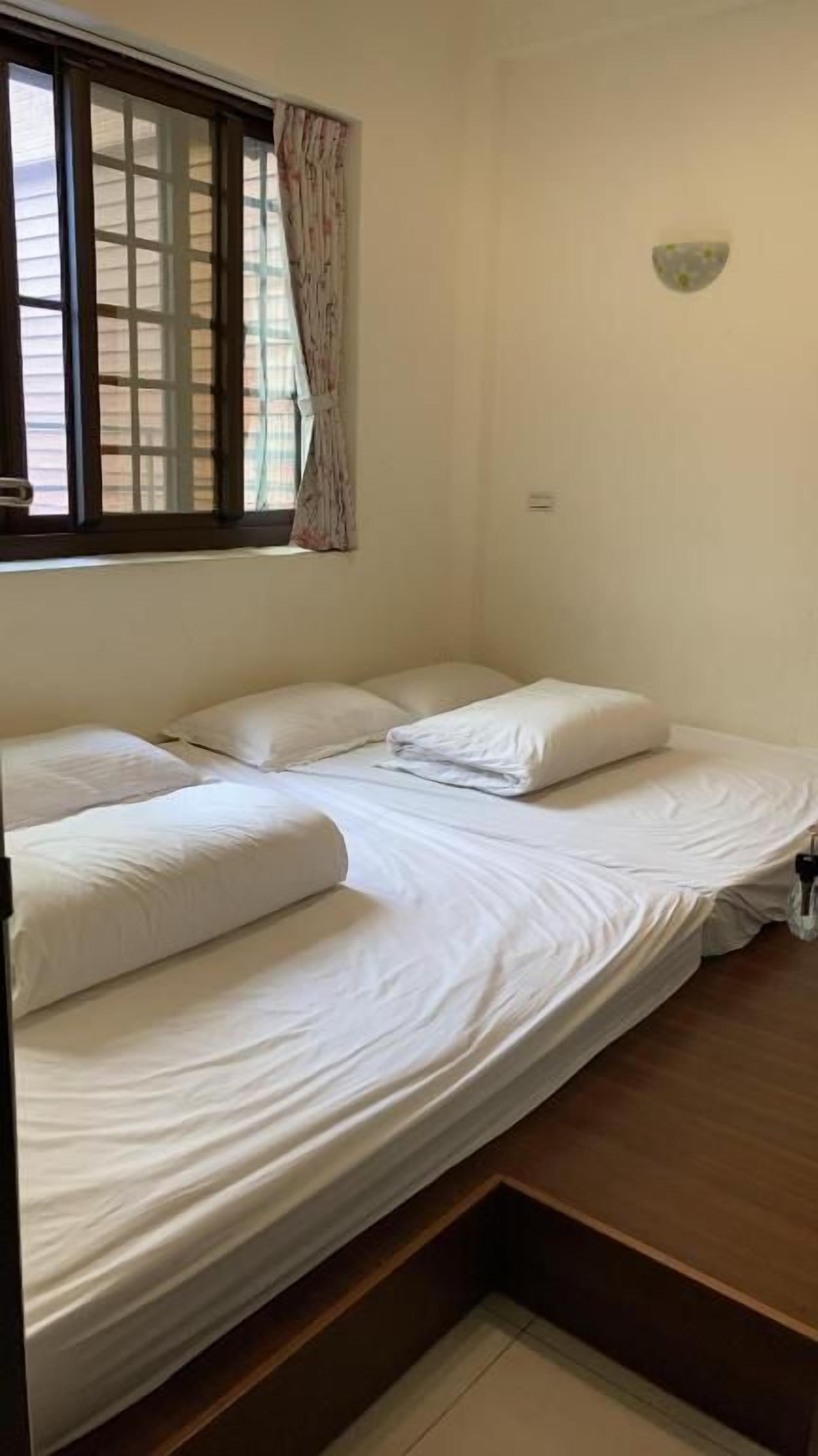 A single-bed room with a wooden floor, a window with curtains, and a wall-mounted light fixture.