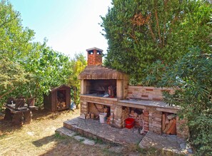 Ein Barbereich aus Ziegelsteinen mit Grill und Schornstein, umgeben von Bäumen und einer kleinen Holzhütte.