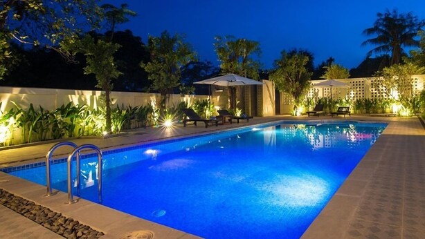 Piscina con sillas de descanso y sombrillas, rodeada de plantas y una valla blanca.