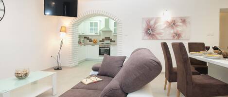 A modern living room with a sofa, a dining table with chairs, a TV mounted on the wall, and a kitchen visible through an archway.