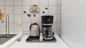 Une cafetière et une bouilloire sont posées sur un plateau dans une cuisine aux carreaux blancs, et une tasse à café est accrochée au mur.