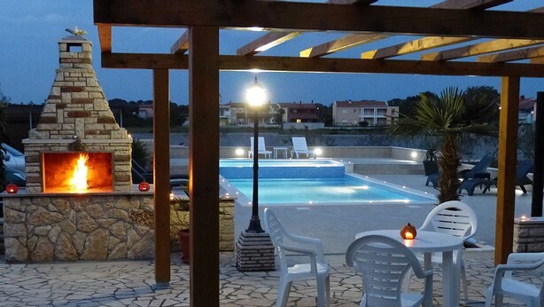 Une cheminée en pierre, une piscine extérieure et des chaises en plastique blanches sous une pergola en bois.