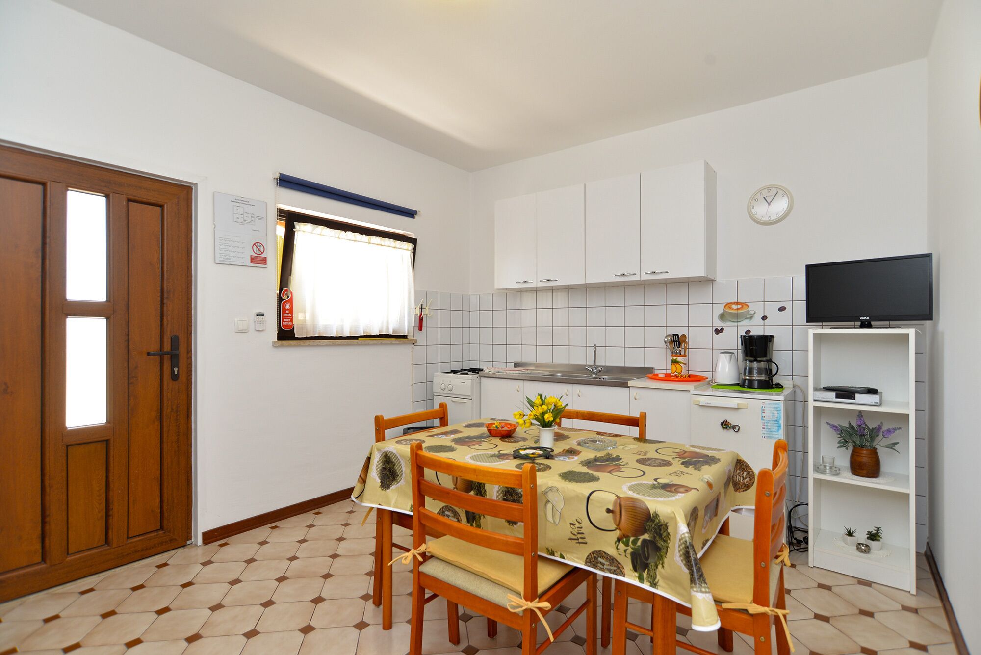 A compact kitchen with white cabinets, a dining table with chairs, a coffee maker, and a television on a shelf.