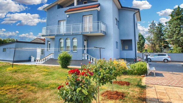 Ein zweistöckiges Gebäude mit Balkon, einem kleinen Garten mit Blumen und einem geparkten Auto auf der Einfahrt.