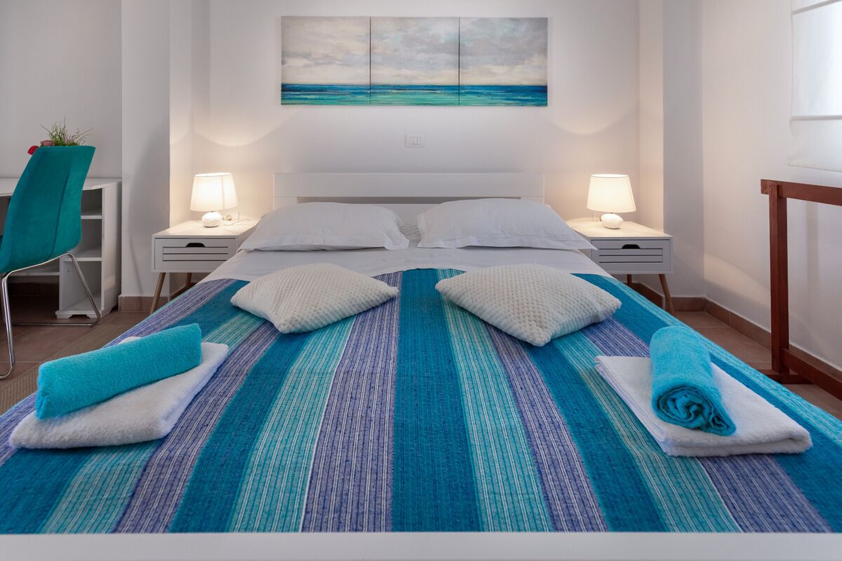 Ein ordentlich bezogenes Bett mit einer blau-weißen, gestreiften Tagesdecke, zwei weißen Kissen und zwei gerollten Handtüchern. Es gibt zwei Nachttische mit Lampen, ein Sessel mit einer Pflanze und ein Bild einer Strandlandschaft über dem Bett.