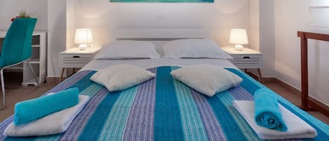 Una cama bien tendida con colcha azul y blanca a rayas, dos almohadas blancas y dos toallas enrolladas. Hay dos mesitas de noche con lámparas, una silla con una planta, y un cuadro con una escena de playa sobre la cama.